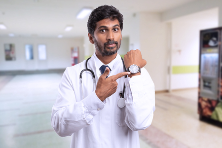 Serious indian male doctor or medic showing wrist-watch with index finger as late for work concept in hospitalの写真素材