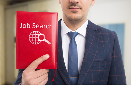 Man wearing suit and tie showing red notebook with job search text and icon with earth and magnifying glass drawing on coverの写真素材