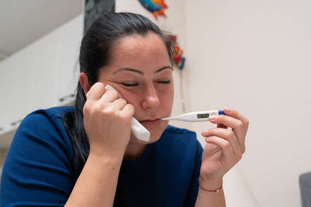 Female model being sick checking body temperature using thermometer as high fever unwell covid19 sars influenza symptom as pandemic conceptの写真素材