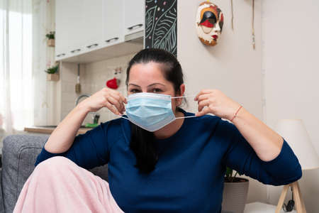 Ill adult woman sitting at home sofa putting on disposable medical or surgical mask to protect from...