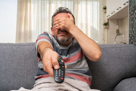 Adult man making scared afraid gesture covering eyes using hands watching horror movie on tv changing channel with remote control as at home pandemic conceptの写真素材