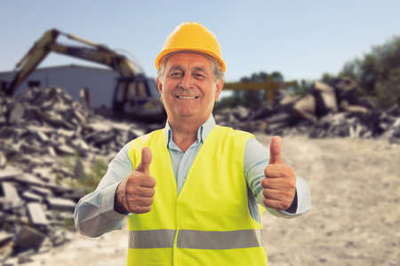Cheerful old male builder making double like gesture using thumbs-up wearing work clothing vest and yellow hardhat on construction site backgroundの写真素材