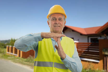 Old male constructor with house background making pause timeout gesture using palms wearing safety hardhat and vestの写真素材