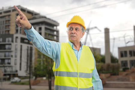 Elderly builder man wearing yellow safety hardhat and work clothing fluorescent vest pointing index finger with building construction site backgroundの写真素材