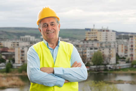 Friendly old male construction worker wearing trades clothing vest and hardhat portrait with crossed arms smiling on town building backgroundの写真素材