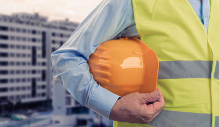 Close-up of male old construction worker holding yellow protective hardhat wearing fluorescent work...