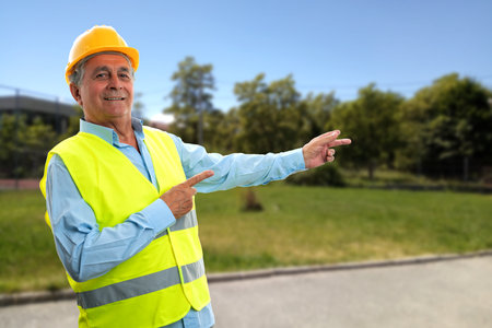 Cheerful elder constructor man smiling pointing index fingers at park nature background wearing fluorescent vest and protective hardhatの写真素材