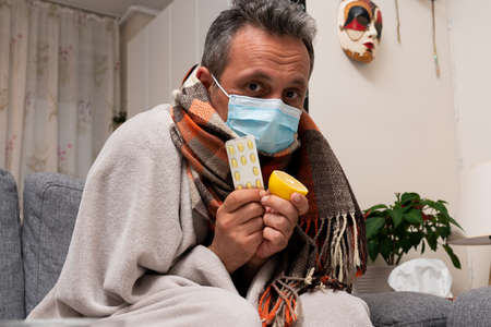 Sick adult male model wearing medical or surgical disposable mask covering neck using colourful scarf presenting lemon for vitaminc immunity and sars covid19 flu cold medical treatmentの写真素材