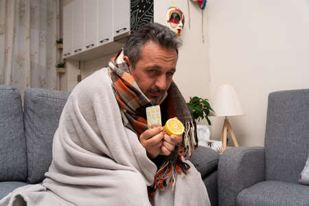 Sick adult man feeling unwell wearing scarf around neck to warm up holding pills as cold sars covid19 pandemic medical treatment and lemon for health immunity covered in blanket at homeの写真素材