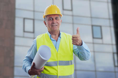 Old male architect contractor wearing yellow hardhat and vest making thumb up like gesture holding...