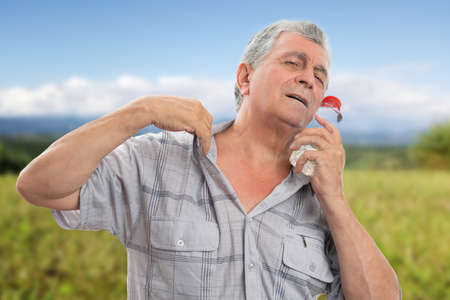 Elderly man wearing casual shirt touching face to cool down using cold water bottle as hot summer fashion concept with nature grass park backgroundの写真素材
