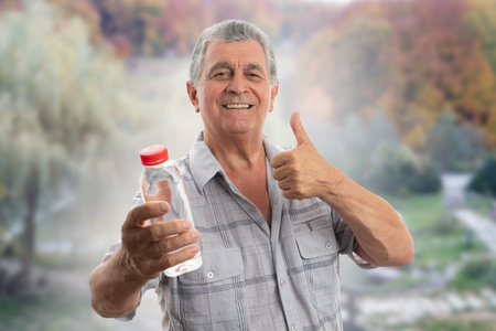 Old man wearing casual summer shirt clothing holding water bottle smiling making thumb-up like gesture as hydration concept forest nature backgroundの写真素材