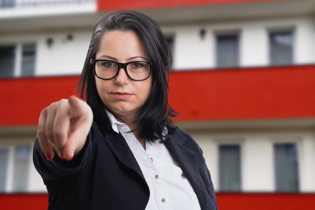 Serious businesswoman wearing spectacles and corporate suit pointing index finger at transparent screen display with apartment building backgroundの写真素材
