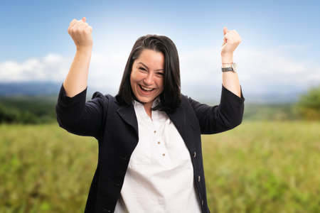 Corporate businesswoman celebrating success making gesture with fists and happy expression smiling cheerful wearing office suit on park field green nature backgroundの写真素材