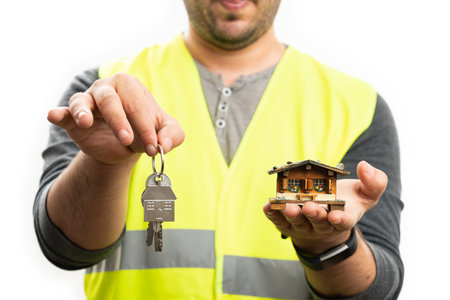Close-up of adult builder man wearing fluorescent vest work clothing presenting miniature wooden house and home keys isolated on white backgroundの写真素材