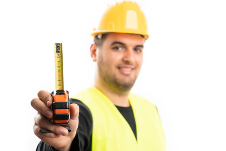 Selective focus close-up of adult builder man in work clothing holding tape measure isolated on...