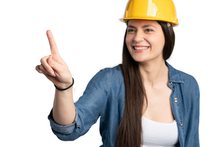 A close-up of a cheerful female construction worker wearing a yellow hard hat and a denim work outfit. She is smiling and pointing upwards, symbolizing innovation, leadership, and forward-thinking in the skilled trades industry.の写真素材