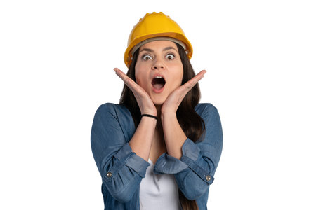 Young female construction worker wearing a yellow hard hat, holding her hands to her face with a surprised expression. She is dressed in a denim shirt, reacting with shock against a white background.の写真素材