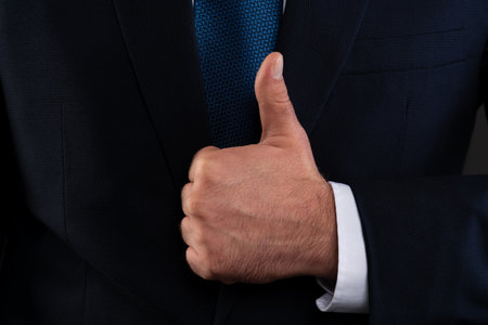 Close-up shot of a man's hand giving a thumbs up gesture while wearing a dark suit and white shirt, symbolizing approval and success in business.の写真素材