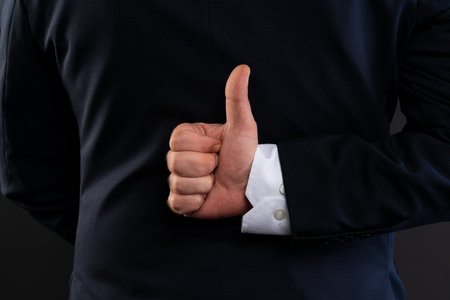 Detailed view of a man's hand making a thumbs up gesture behind his back, dressed in formal business...