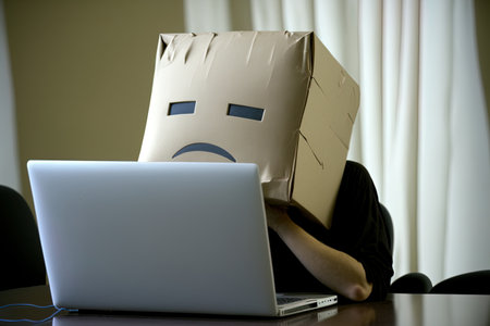 The topic of stress and privacy concerns on the internet is represented by a depressed person working on a laptop while wearing a paper box over his headの素材