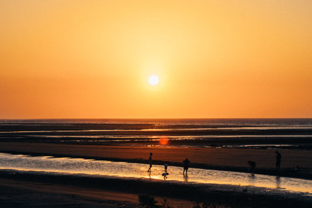 Sunset on the beach with people walking in the foreground and the sun reflecting in the waterの写真素材
