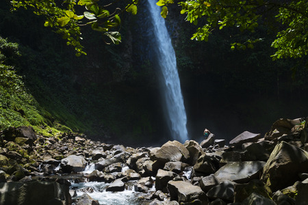 View of the La Fortuna Waterfall with one man standing on a rock in Costa Rica, Central America; Concept for travel in Costa Ricaの写真素材