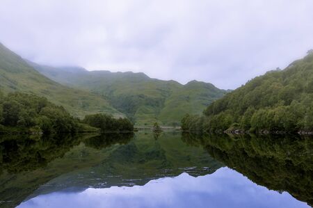 Misty and serene landscape of a lake and mountains in the Highlands of Scotland, United Kingdom;...