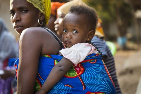 Bissau, Republic of Guinea-Bissau - January 29, 2018: Portrait of a baby daughter being carried on her mother shoulders, at the Bissaque neighborhood in the city of Bissau, Guinea Bissau.のeditorial素材