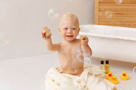 Bathing the baby in white bath with yellow rubber ducks. Kid sitting next to bath and smiling. A lot of soap bubbles flying in the bathroom.の写真素材