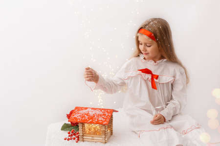 Little blonde girl playing with wooden doll house with lights in the middle. Little kid in nightgown is sitting on table and pours snow on the red roof of the house Holiday concept.の写真素材