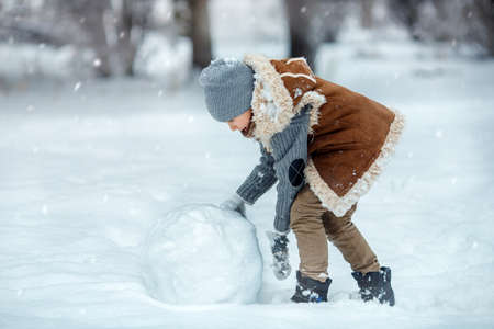 Happy boy playes on snowy winter day in the park. Child playing with snow in winter and rolling snow ball for making a snowmanの写真素材