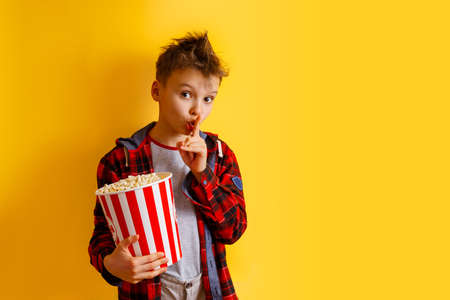 Cute boy who is asking to be quiet with finger on lips with huge popcorn bucket on yellow background. Kid is wearing casual plaid shirts outfit isolated. Movie concept. Teenager showing quiet gestureの写真素材