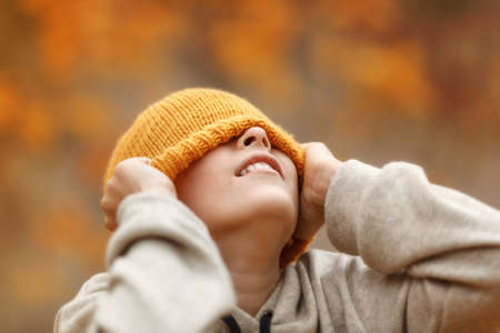 Boy is hiding his eyes by ginger knit hat in the autumn park. Bright colour background. Close up portrait of a cheerful boy.の写真素材