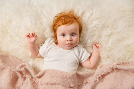 Red hair baby girl is lying at home under the blanket. Kid is looking at camera. Cheerful toddler