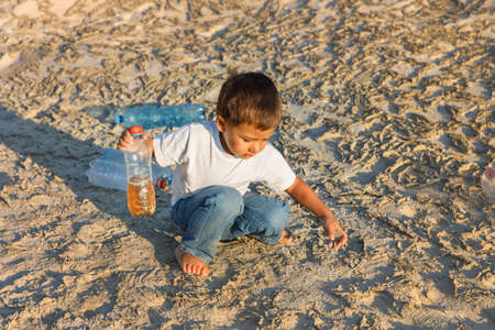 Sad kid drinking water on crack ground. Concept drought and shortage of water crisis. Thirsty kid sits and holds dirty water from a bottle on dry sand. Earth day.の写真素材