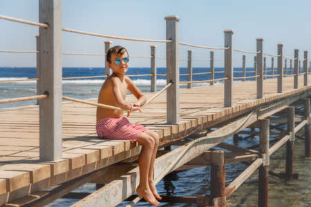 The kid sits on the pier against the sea, the boy wearing sunglasses and sitting on top of the sea...