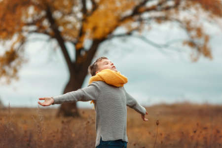 Boy in grey sweater and yellow scarf on a field in autumn day. Kid raising hands over grey stormy sky background and beautiful tree.. Fresh air, environment conceptの写真素材