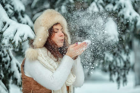 A women in a sweater and hat in winter blowing snow off her hands in a snowy forest. Funny female...