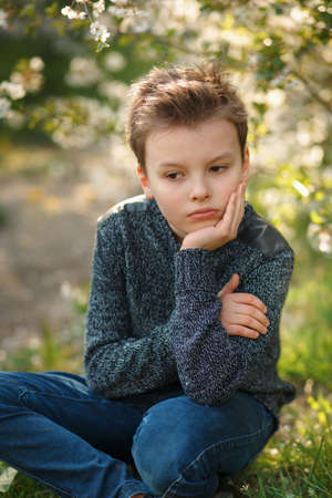 Boy is sitting on the grass in the blossom garden on beautiful spring day. Kid is sad prop one's chin with one's handの写真素材