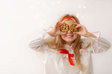 Blonde girl in nightgown hidding her eyes behind cookies. Little kid is sitting on table with miniature house and bottle of milk and wondering. Holiday concept.の写真素材
