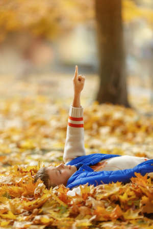 Happy boy in sweater and blue vest laying on autumn leaves and and shows the index finger up. autumn holiday time. Pointer fingerの写真素材