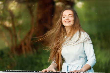 Girl playing a synthesizer piano in nature at sunset. A woman is 22 years old. An electric piano stands in a field in a village. Her hair is blowing in the windの写真素材