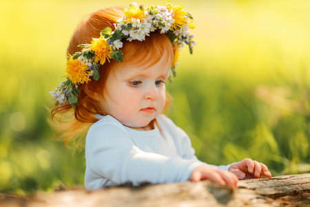 Portrait of a cute little girl is wearing beautiful spring wreath outdoors, space for text. Red hair baby is spending time on nature. Thousand baby.の写真素材