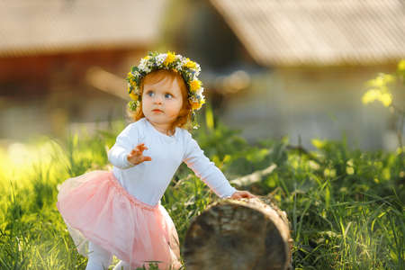 Cute little girl is wearing beautiful spring wreath outdoors, space for text. Red hair baby is spending time on nature, she standing and hold on to a stumpの写真素材