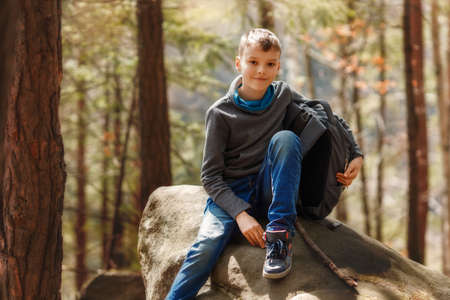 A boy sits on a large stone in nature. Kid is smiling and looking at camera, backpack is near the...