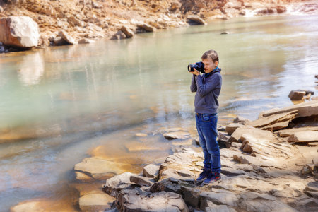 Photographer boy near a mountain river standing on the rock. Kid enjoying the moment of the beautiful blue water in sunny day. Boy is holding photo camera and taking photo of the view.の写真素材