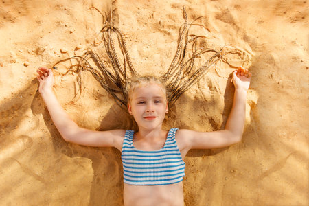 A smiling girl with braids lies on the sand in a striped swimsuit, happy and bathed in sunlight.の写真素材