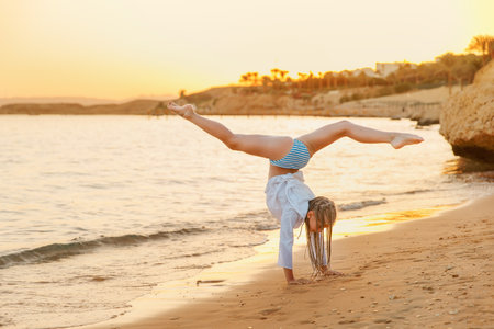 A girl is doing acrobatic exercises on the beach at sunset. She is standing on his hands in the...