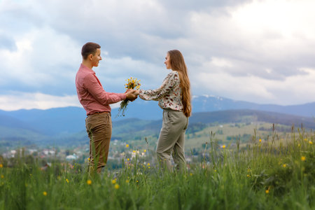 A boy and girl stand in a meadow, facing each other and holding hands. A bouquet of wildflowers is...